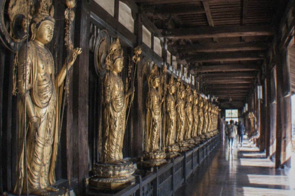 A long wooden temple hall interior lined with rows of gilded standing statues receding into the distance