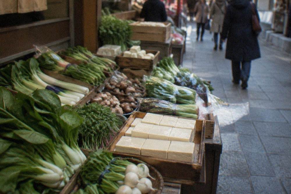 Fresh green vegetables and tofu blocks displayed on wooden trays at a Japanese market stall