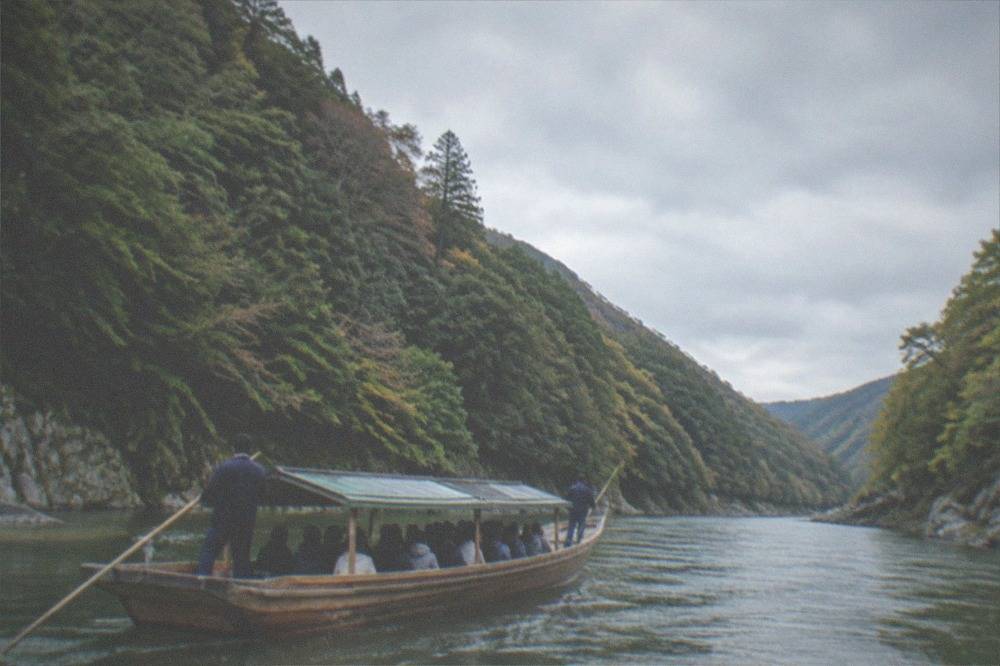 A long wooden river boat carrying passengers through a narrow green gorge with boatmen standing at bow and stern