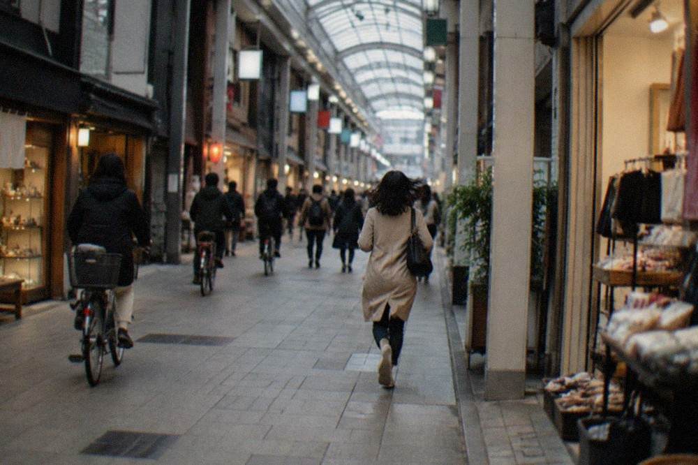 A covered shopping arcade in Kyoto with lit shopfronts, bicycle traffic and pedestrians under a glass roof