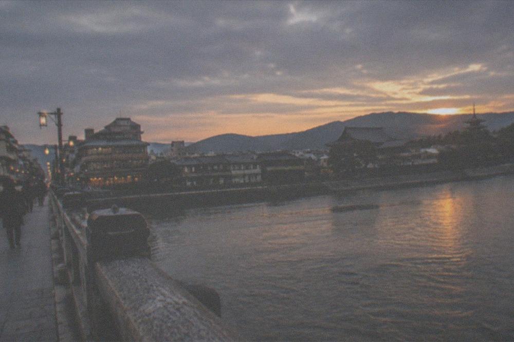 A stone bridge over a wide river in Kyoto at sunset, with the eastern hills and temple rooftops beyond