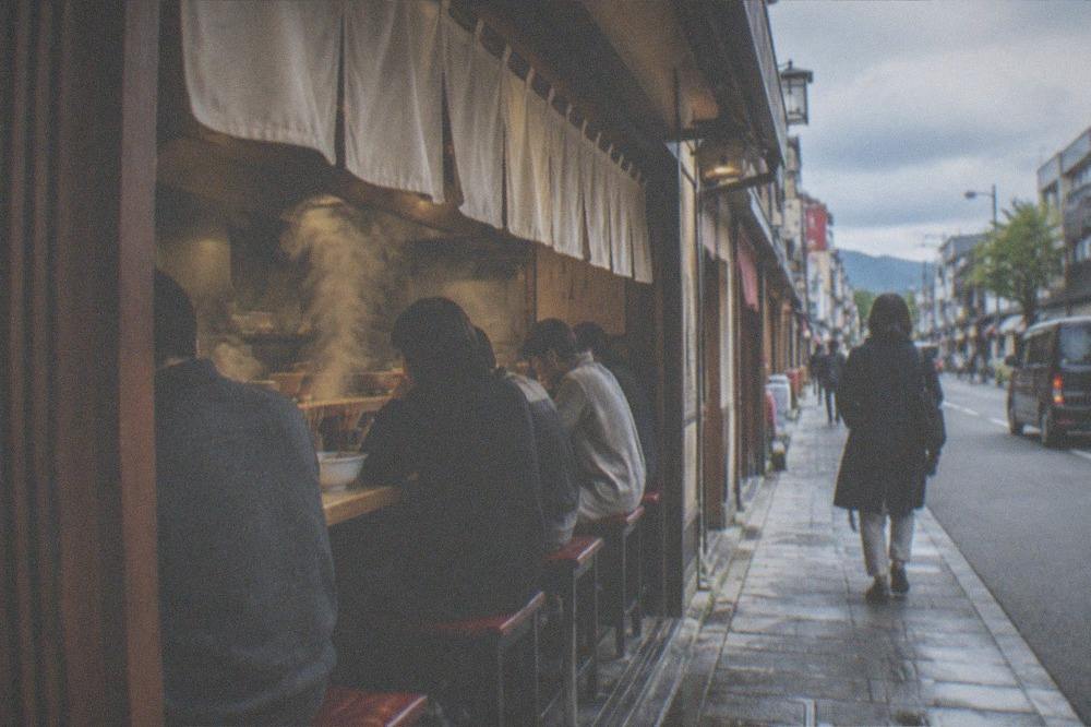 A narrow ramen counter restaurant with steam rising from bowls and customers seated side by side