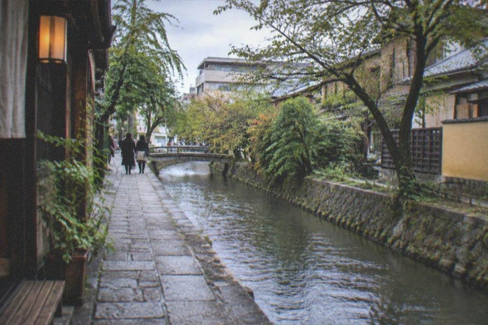 A quiet stone path beside a narrow canal running between trees and traditional houses