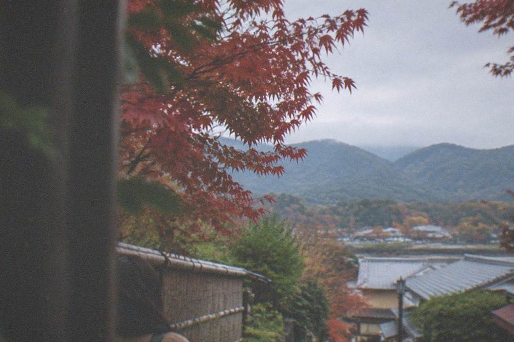 Red maple leaves framing a view over rooftops toward the mountains around Arashiyama