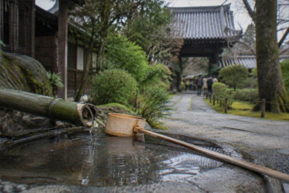 A low stone water basin with a bamboo ladle beside a mossy temple path