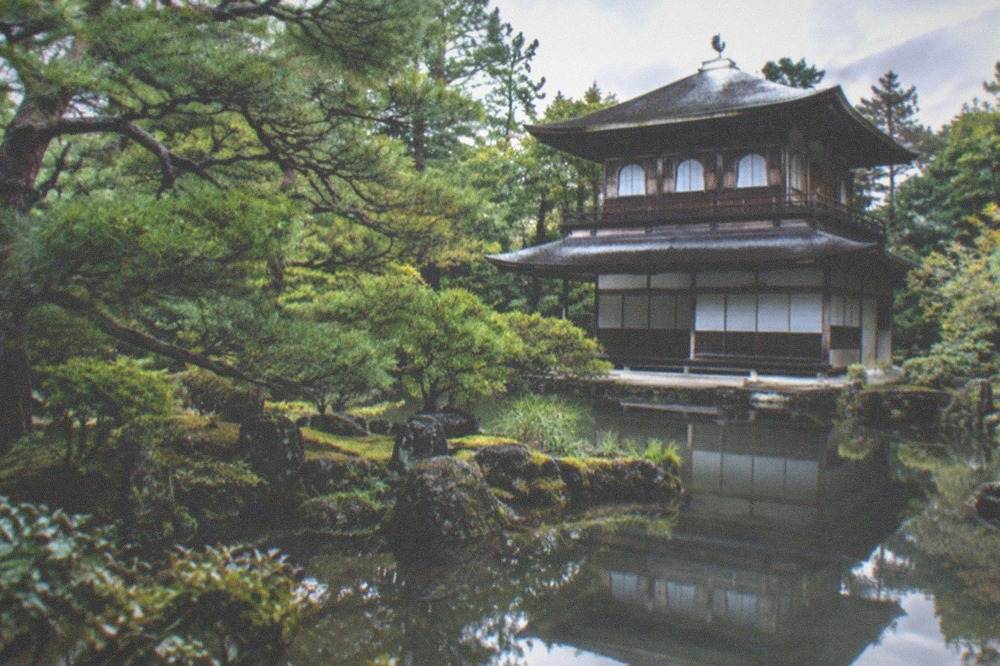 A weathered two-storey pavilion reflected in still water surrounded by trees
