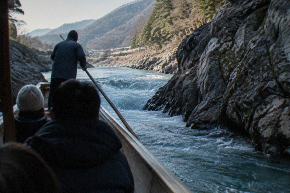 A boatman steering a wooden river boat through a small rapid between rock walls