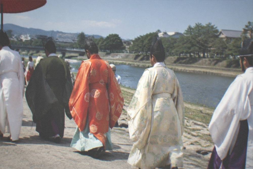 A procession of costumed participants crossing open ground beside a river in daylight