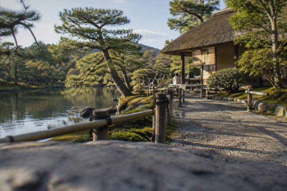 A manicured Japanese garden path beside a pond with pine trees and a thatched teahouse in Kyoto