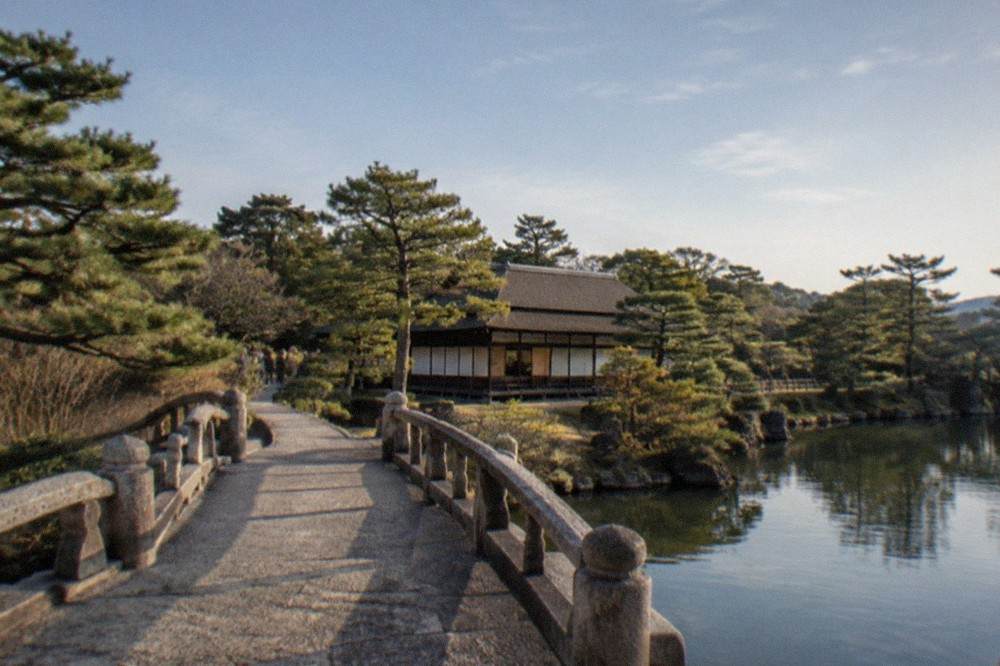 A stone bridge crossing a garden pond toward a teahouse surrounded by pines in Kyoto