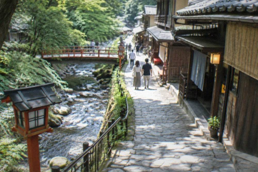 A quiet mountain village road running beside a stream, with traditional wooden buildings and forest slopes on both sides