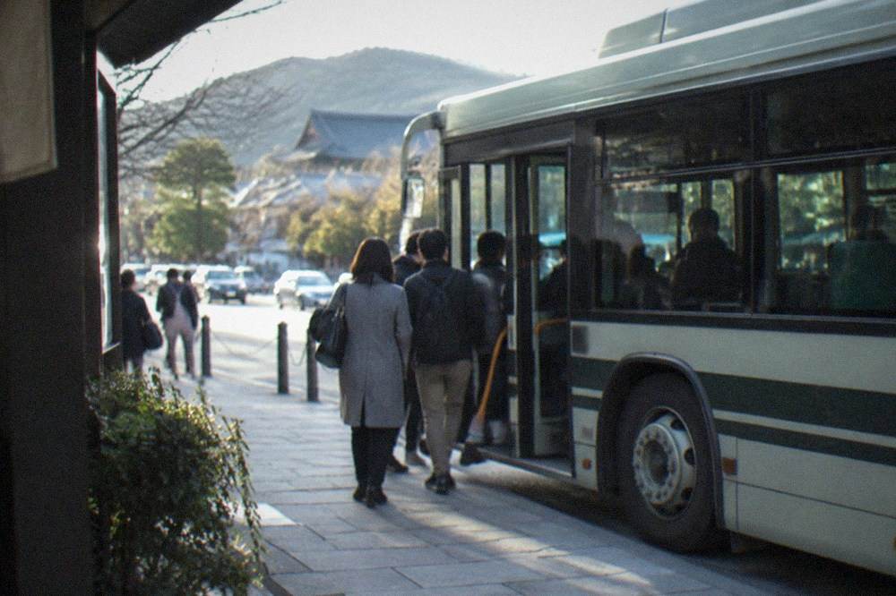 A city bus stopped at a busy street corner in Kyoto with passengers boarding