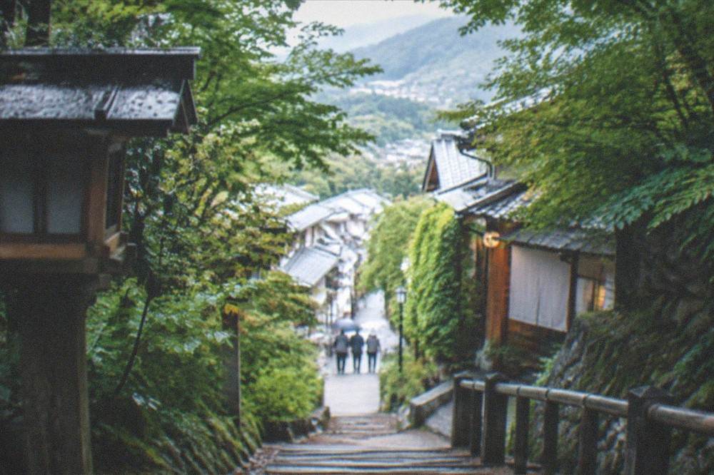 A forested mountain trail with wooden steps leading toward a village