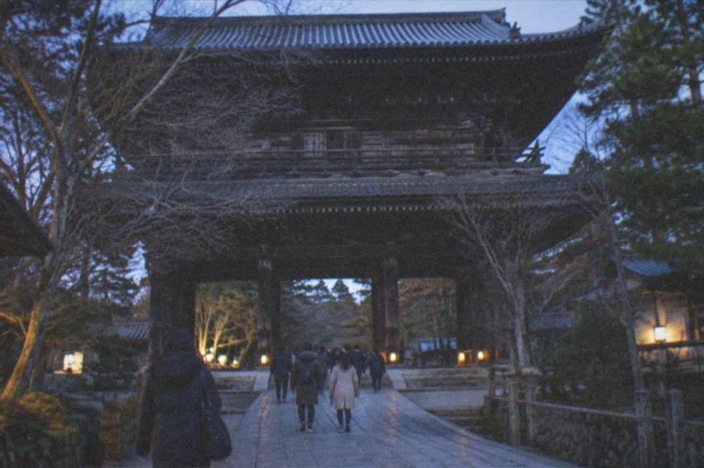 A large wooden temple gate in winter with bare trees and soft low light on the stone approach