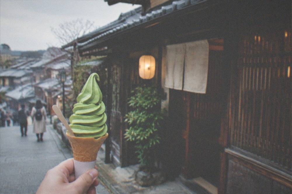 A matcha soft serve ice cream cone held in front of a wooden Kyoto teahouse facade