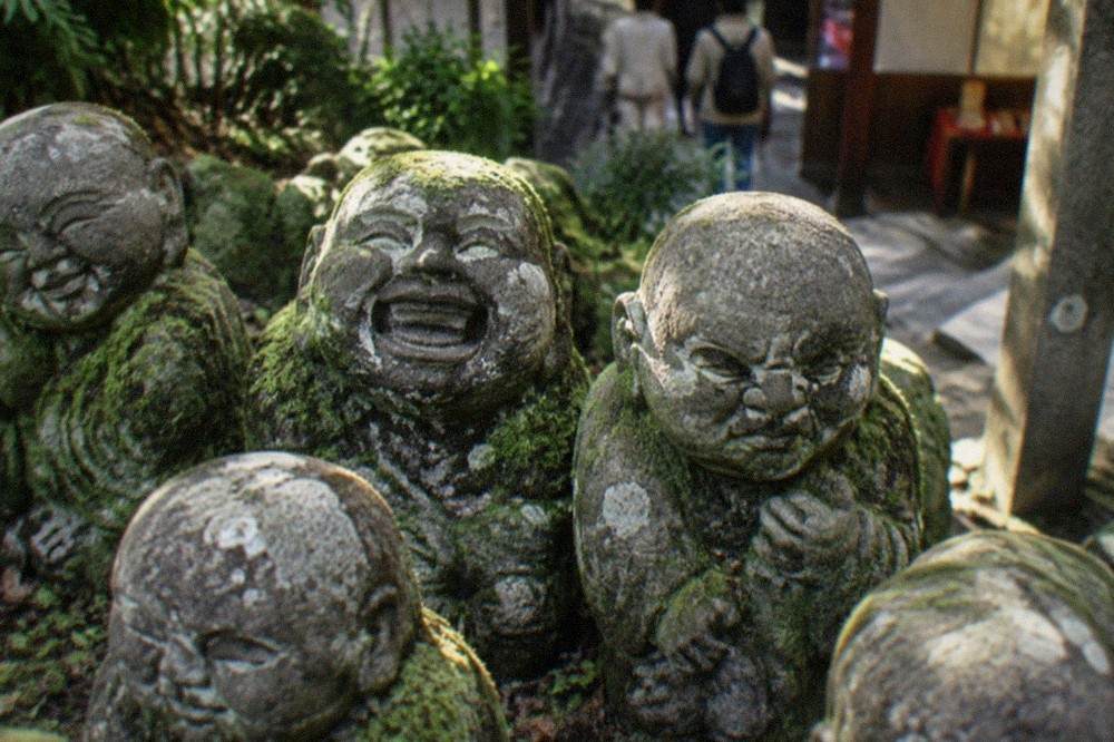 Close view of several weathered stone rakan figures with different facial expressions, moss on their shoulders