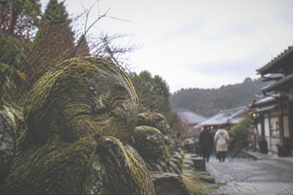 A weathered stone figure with a cheerful carved face partly covered in green moss