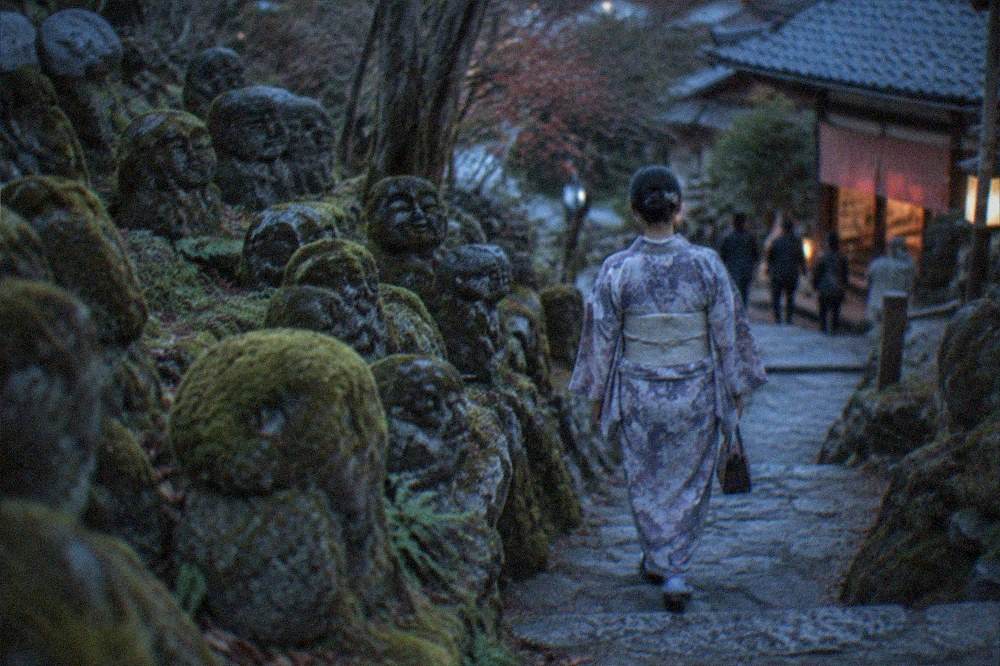 A visitor in a kimono walking a stone path between mossy stone figures on a temple hillside