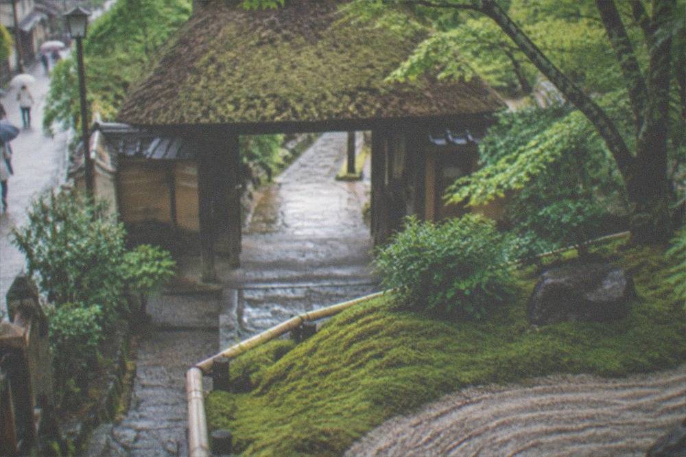 A thatched temple gate behind a moss bank with raked sand patterns