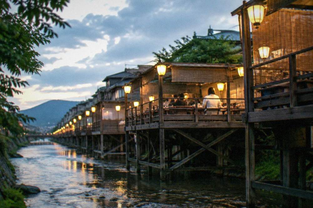 Wooden summer dining platforms extending over a shallow river at twilight with lanterns glowing