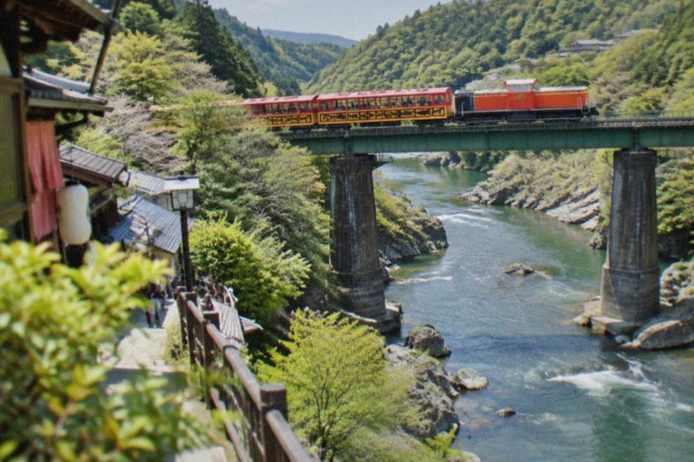 A vintage sightseeing train in bright colours crossing a bridge above a forested river gorge in Arashiyama