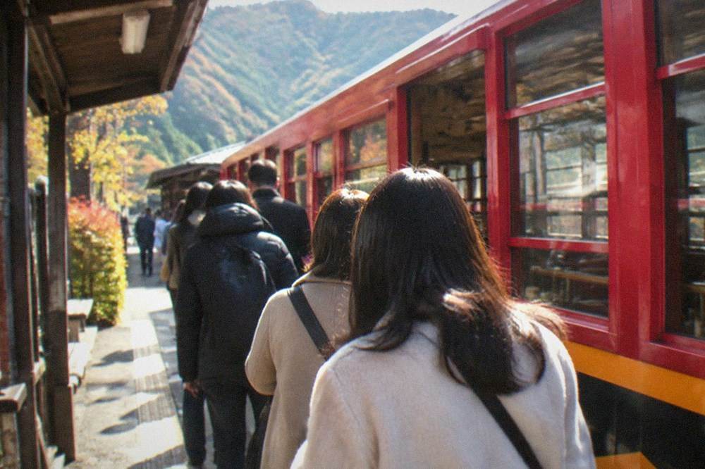 Travellers queueing at a small rural station platform beside a red sightseeing carriage