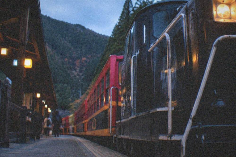 A vintage sightseeing train at a small wooden station platform with forested hills behind