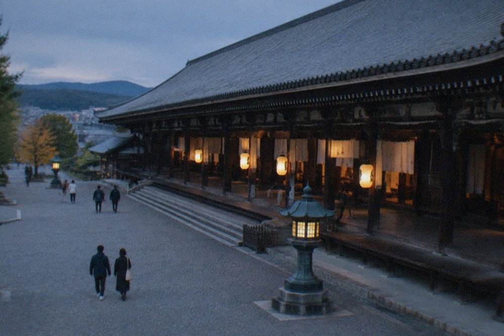 "A long wooden temple hall viewed from its gravel forecourt, with a tiled roof and lanterns hanging at the entrance