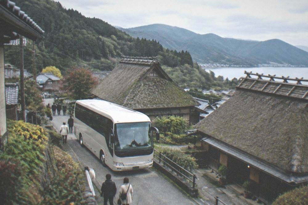 A tour bus parked on a narrow road beside thatched-roof farmhouses in a mountain valley