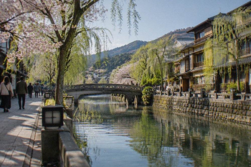A willow-lined canal running between wooden ryokan buildings in a Japanese hot spring town