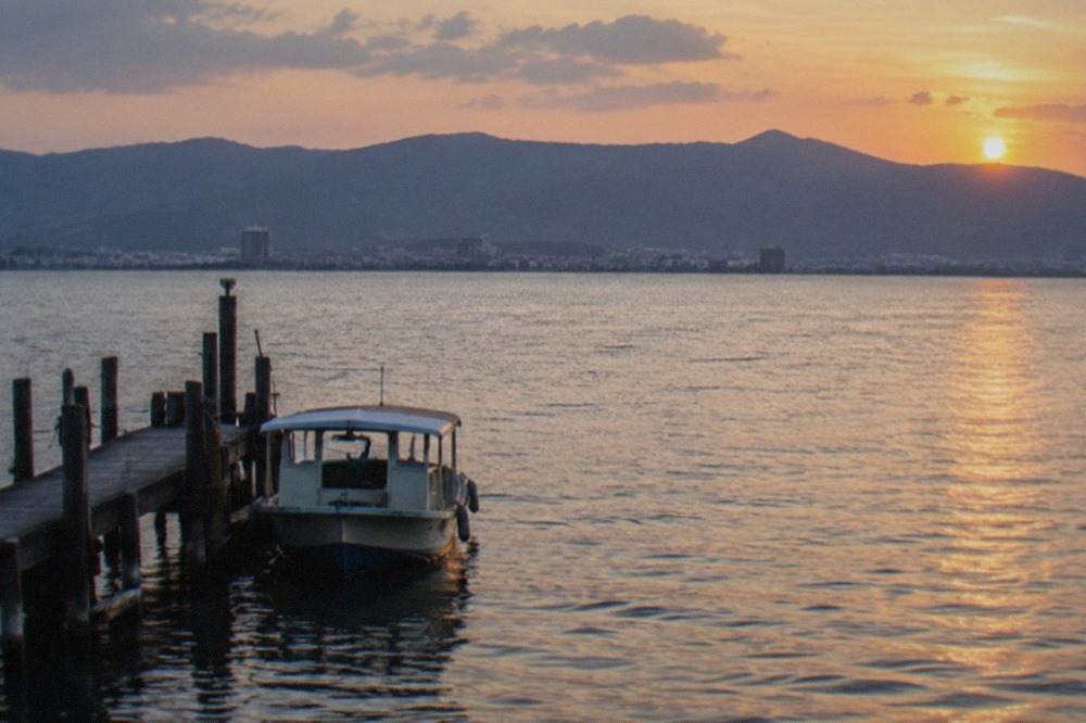 A wide lake at dusk with mountains on the far shore and a small boat near a wooden pier