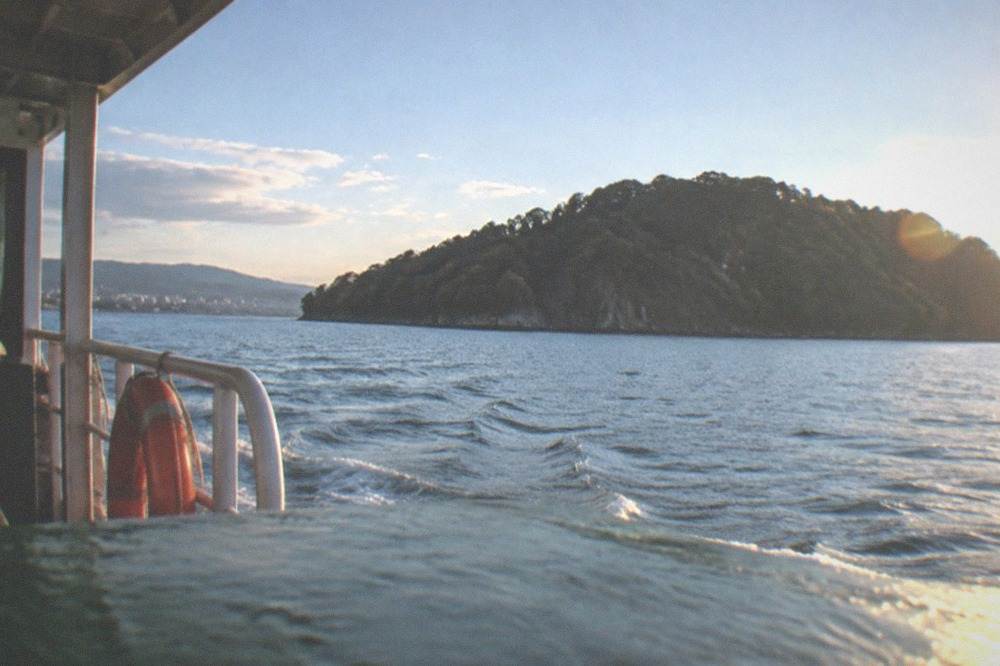 A small ferry crossing open water toward a forested island under a blue sky
