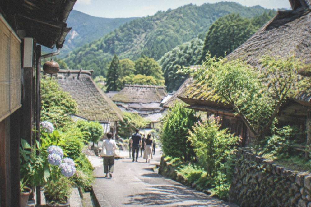 A village lane between thatched farmhouses with green mountains rising behind the rooftops