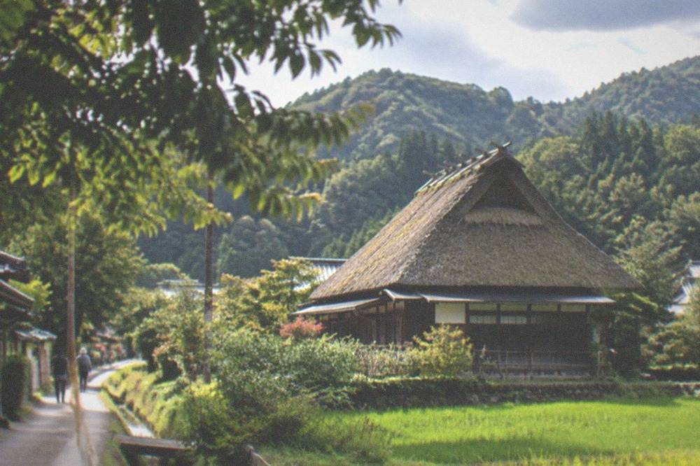 A thatched farmhouse surrounded by green fields and wooded hills in the Kyoto countryside