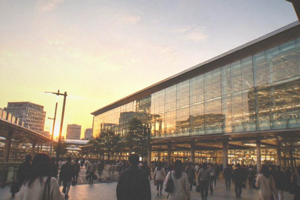 The glass facade of a large railway terminal with streams of passengers at dusk