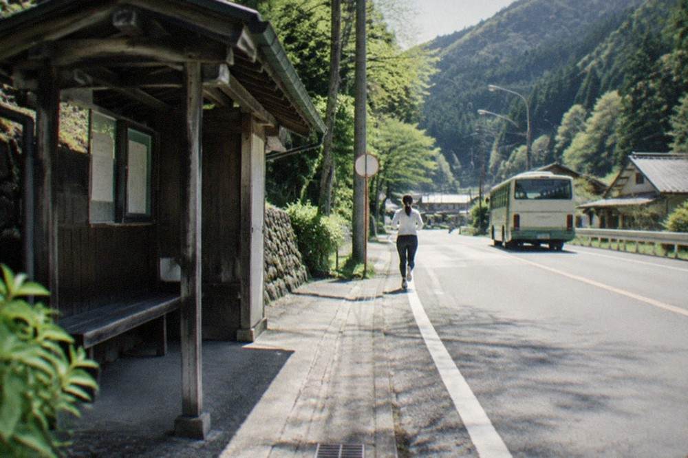 A rural bus stop with a simple shelter beside a forested mountain road