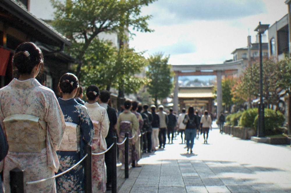 Spectators in kimono waiting behind a low barrier along a shrine approach path