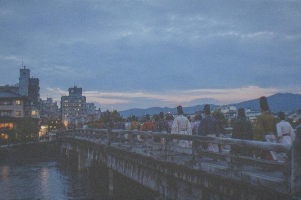 Procession participants in layered court robes crossing a stone bridge over a river
