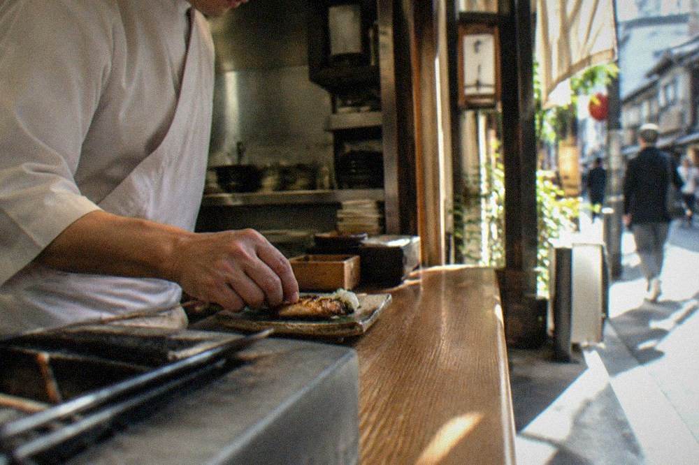 A chef placing a small grilled course onto a counter in a narrow Kyoto restaurant kitchen