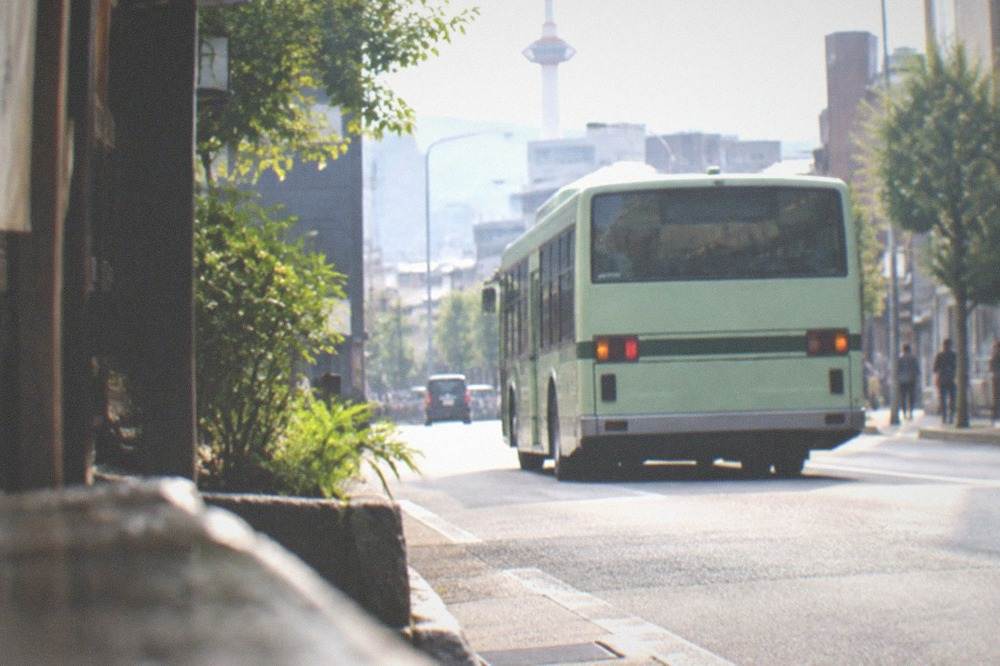 A green city bus pulling away from a stop on a narrow Kyoto street