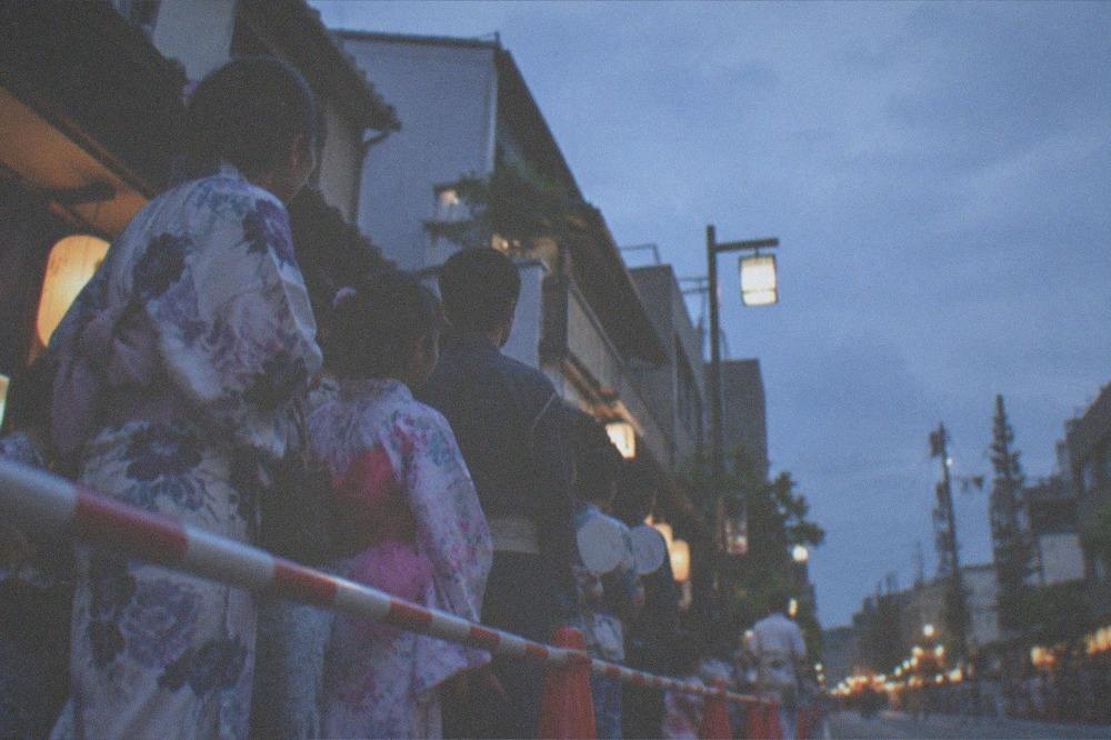 Families in yukata waiting behind a barrier along a parade route in the morning