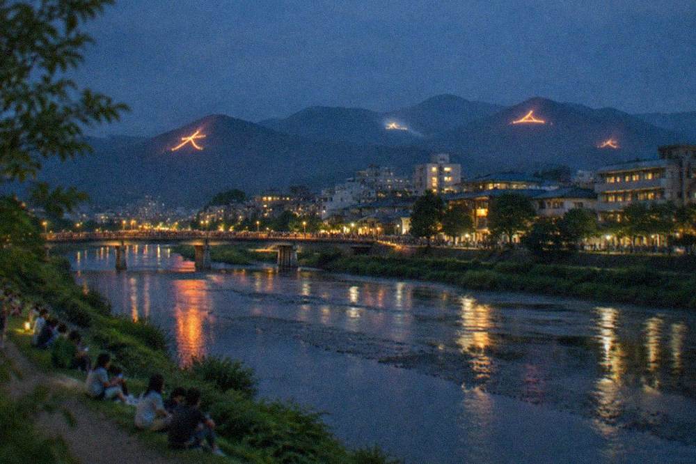 A wide night view of Kyoto with multiple fire shapes glowing on dark mountains around the city