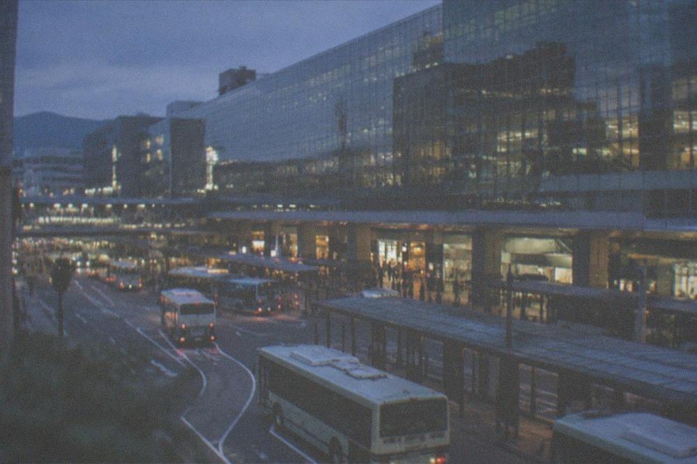 The tall glass facade of a large station building seen from a plaza with buses below