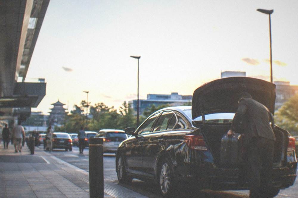 A driver loading suitcases into the boot of a black sedan outside a hotel