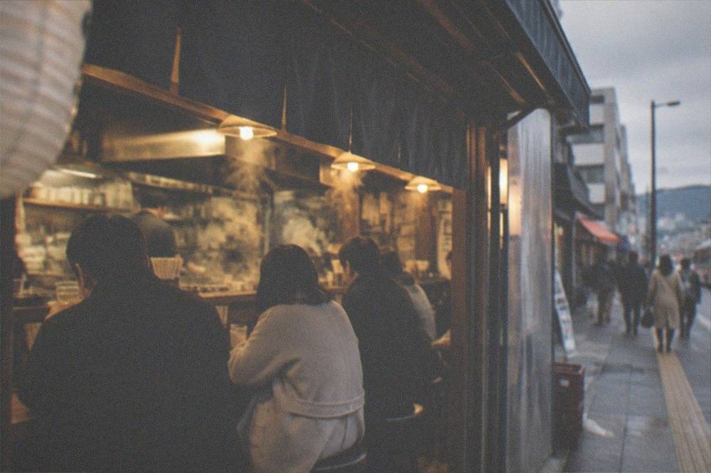 Customers seated at a busy izakaya counter with steam rising from the kitchen