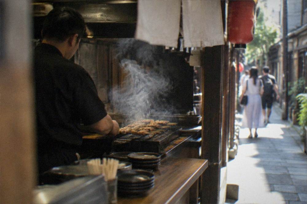 A chef grilling skewers behind a small izakaya counter with smoke rising