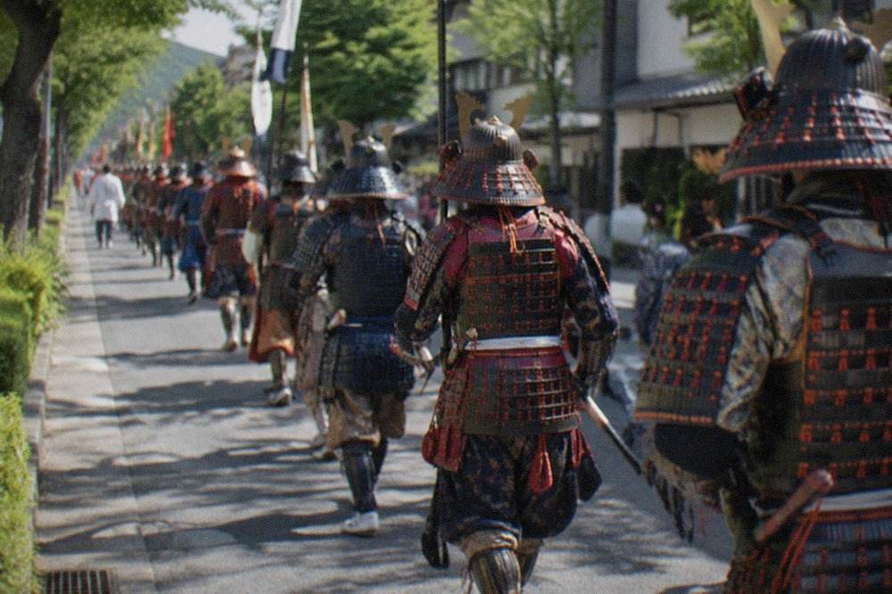 Costumed marchers in historical armour walking in a long procession along a tree-lined Kyoto street