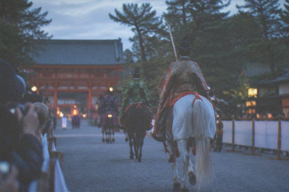 Spectators with cameras pressed along a barricade as costumed horsemen ride past in Kyoto