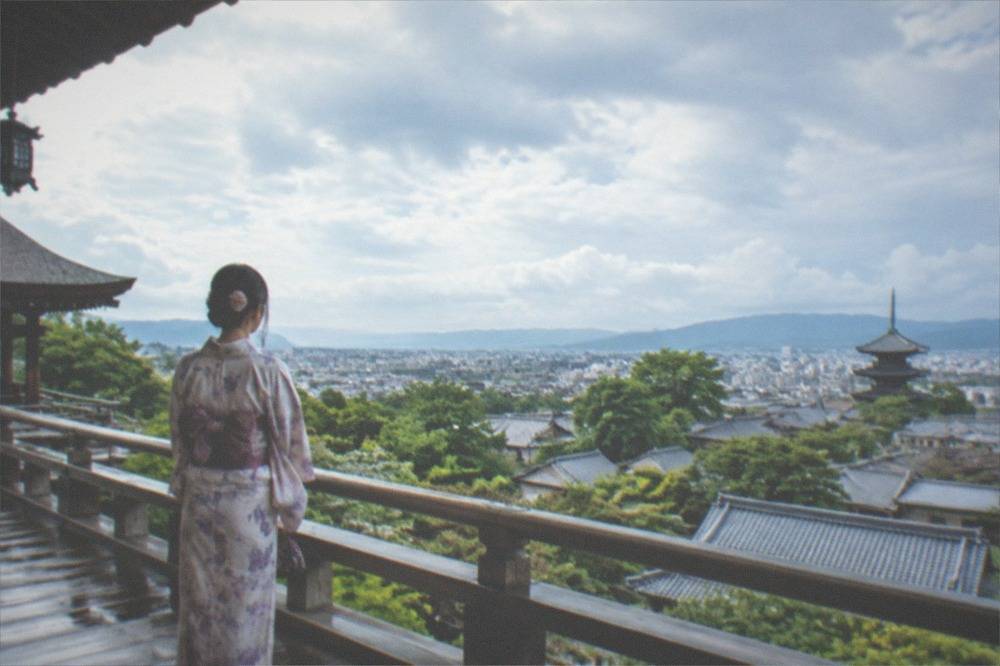 A visitor in kimono standing on a wooden temple veranda overlooking Kyoto rooftops