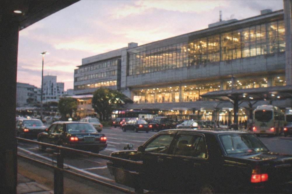 The illuminated facade of a large station building at dusk with a taxi rank in the foreground